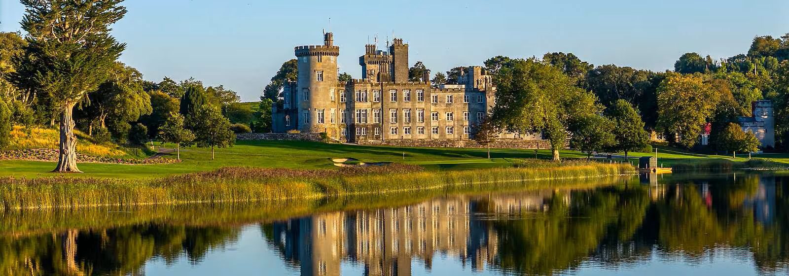 A large stone castle with multiple towers sits on a green lawn beside a calm lake, with the Sunflower Children’s Hospice and surrounding trees reflected in the water under a clear blue sky.