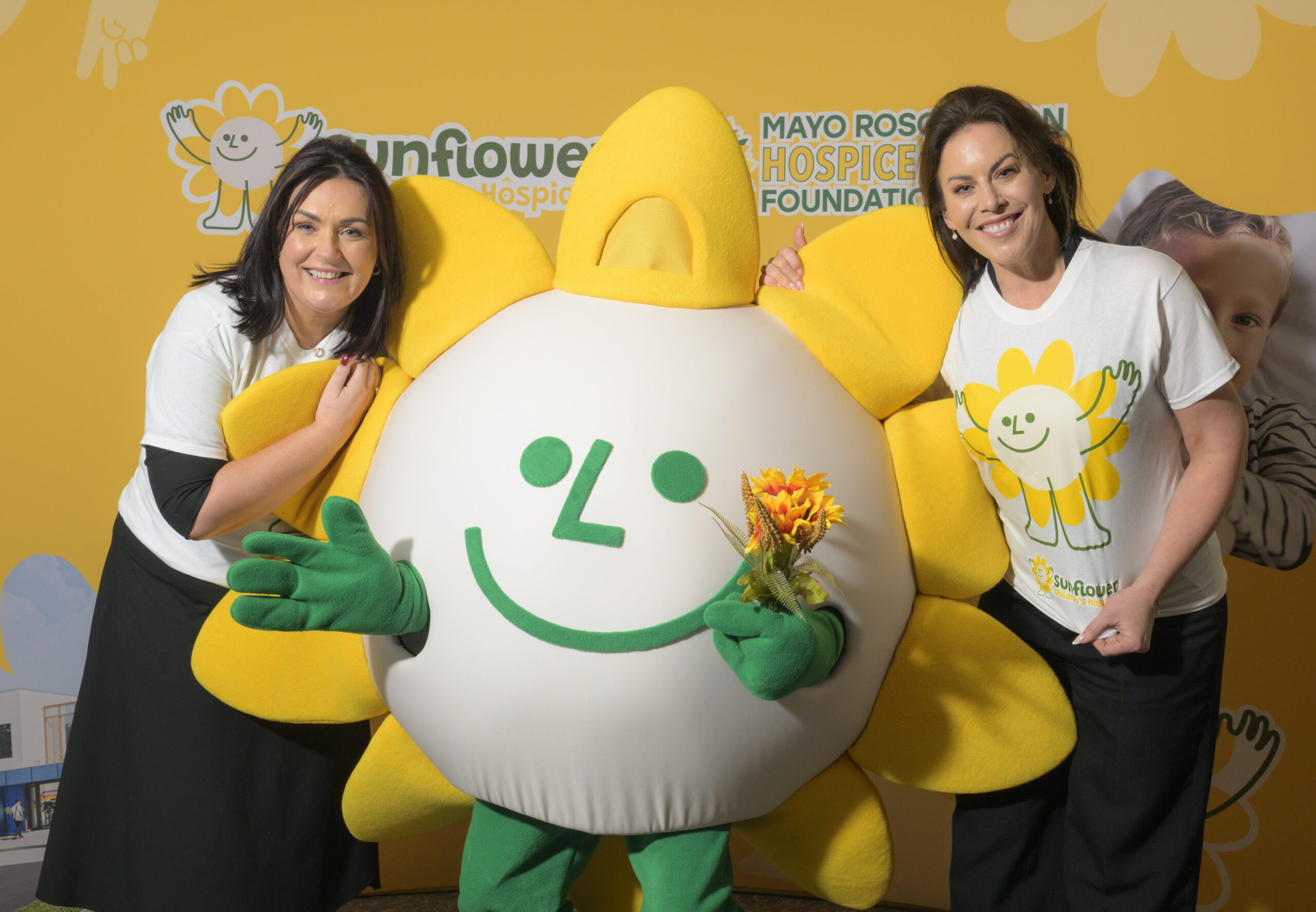 Two women stand beside a person in a large sunflower mascot costume holding flowers, all smiling at the camera. The background features bright yellow sunflower-themed decorations and Sunflower Children’s Hospice logos.