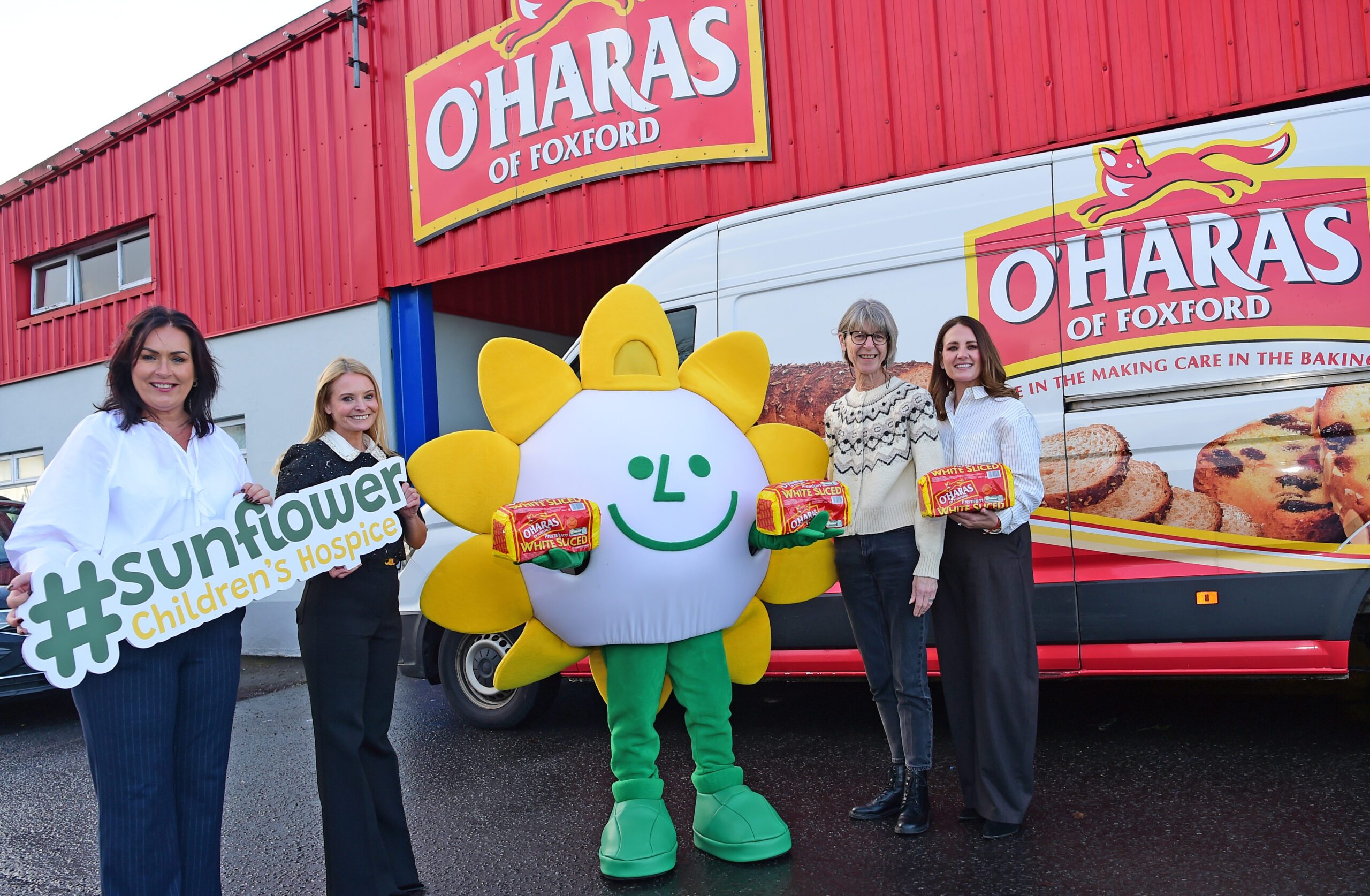 Four women stand outside an O'Haras of Foxford van with a person in a sunflower mascot costume. Two women hold Sunflower Children’s Hospice signs, whilst the others display O'Hara’s baked goods in support of Sunflower Children’s Hospice.