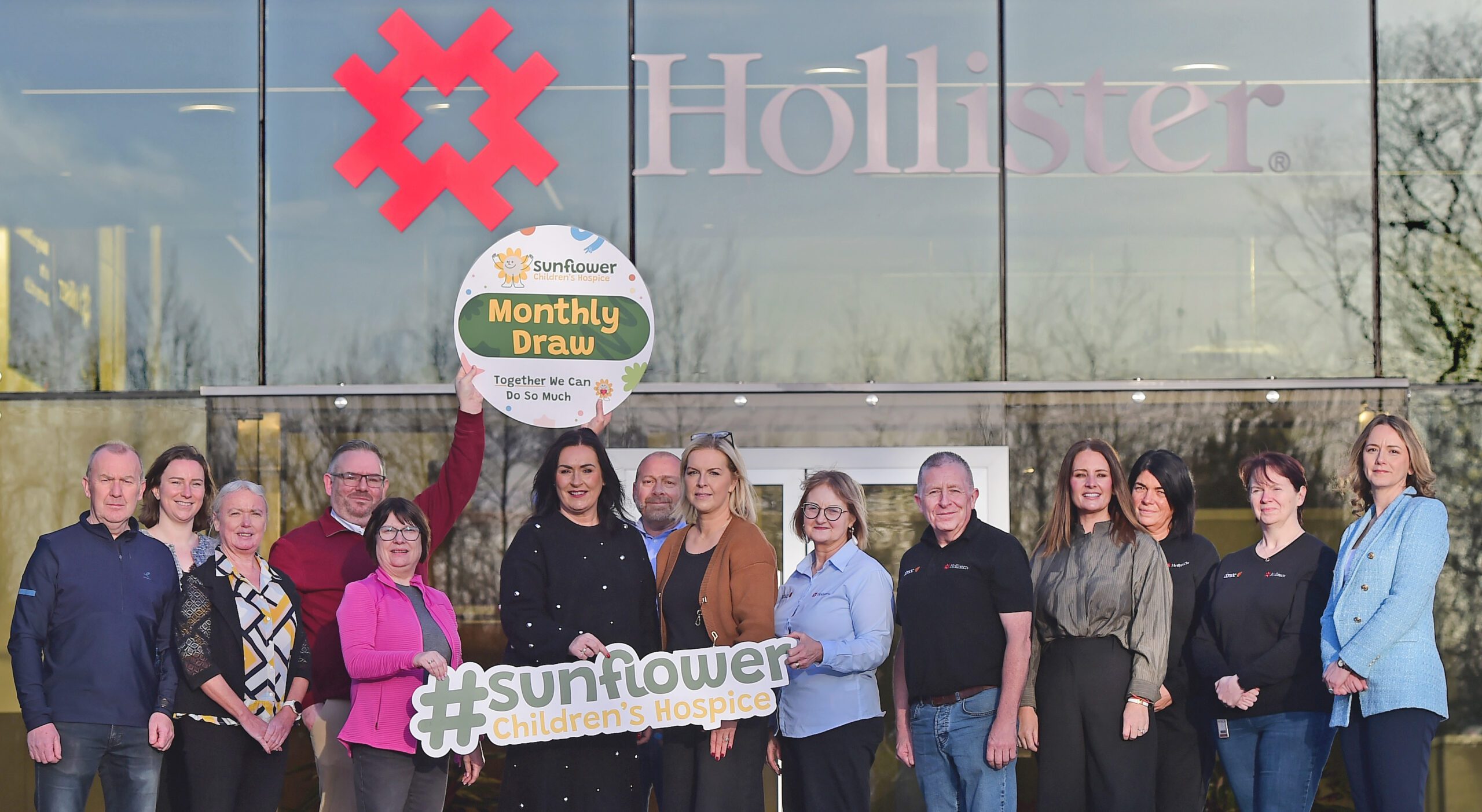 A group of people stand in front of a building with a Hollister sign, holding Sunflower Children's Hospice signs and promoting a monthly draw. The group smiles and poses for the photo.