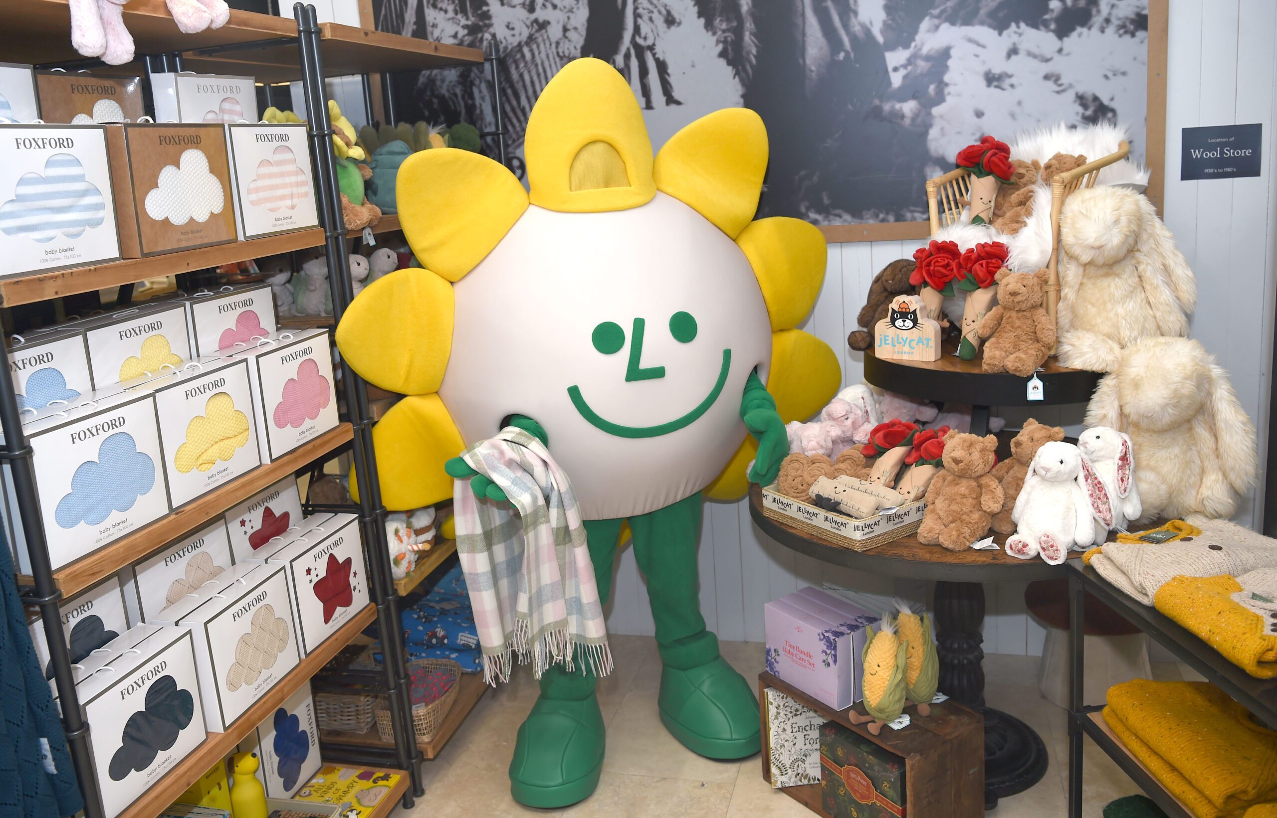 A person in a large, cheerful sunflower costume stands in a cosy shop, holding a scarf. The shop, supporting Sunflower Children’s Hospice, displays soft toys, wool, and colourful scarves on shelves and tables.