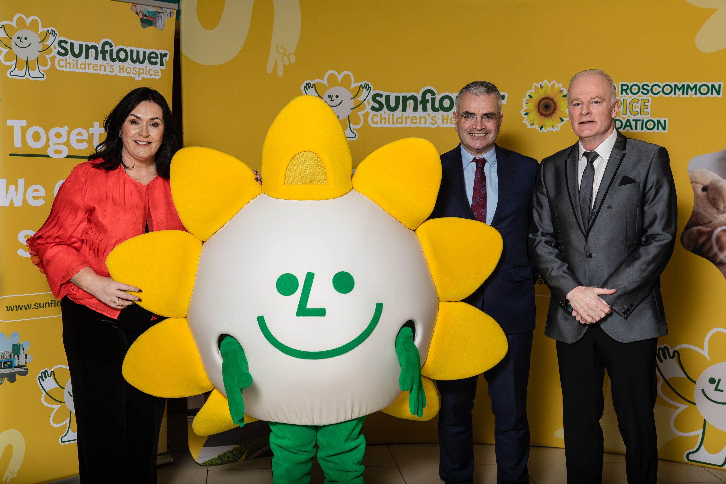 Three adults stand beside a cheerful sunflower mascot, all smiling and dressed formally. Sunflower Children’s and TELUS Digital banners are visible in the yellow-themed background, highlighting their Bound for Canada partnership.