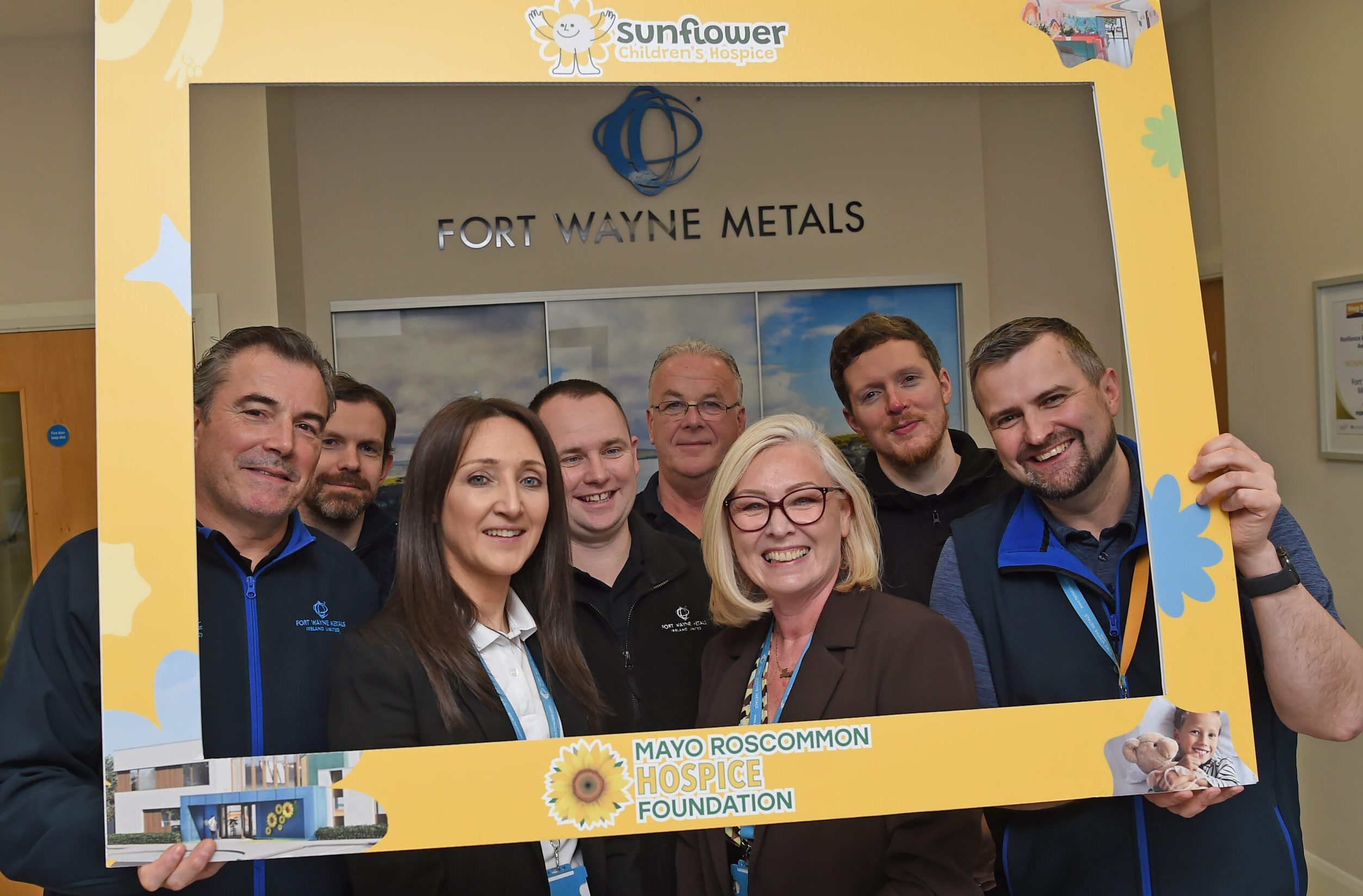 A group of eight smiling people pose together inside a building, holding a large yellow Sunflower Children’s Hospice frame with a Fort Wayne Metals sign on the wall behind them.
