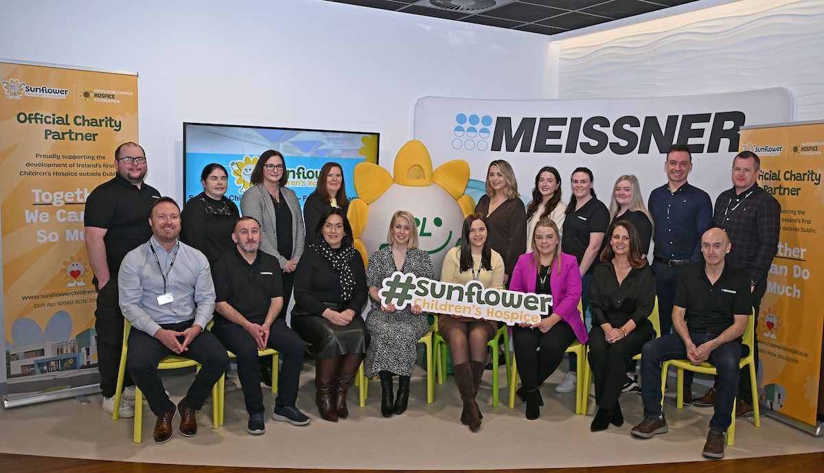 A group of adults poses for a photo indoors with the Sunflower Children’s Hospice mascot, banners for Meissner and Sunflower Children’s Hospice, and a large #sunflower Children's Hospice sign. Some are seated whilst others stand behind.