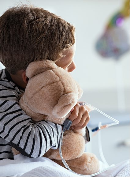 A young child wearing a striped shirt hugs a plush teddy bear whilst sitting in a hospital bed at Sunflower Children's Hospice, with a drip attached to their hand. The atmosphere appears comforting despite the medical setting.