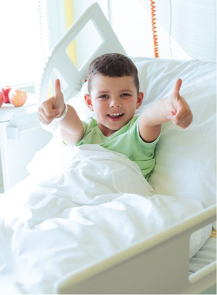 A young boy lies in a hospital bed at Sunflower Children’s Hospice, smiling and giving two thumbs up. He appears cheerful, wearing a light green shirt with a hospital wristband, and is partially covered by a white blanket.