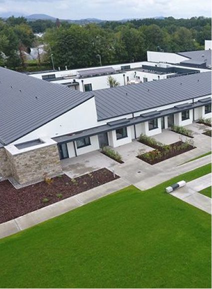 A modern building with white walls and a dark grey roof, featuring large windows and stone accents, sits surrounded by green lawns and landscaping—Sunflower Children’s Hospice provides a peaceful retreat under a cloudy sky.