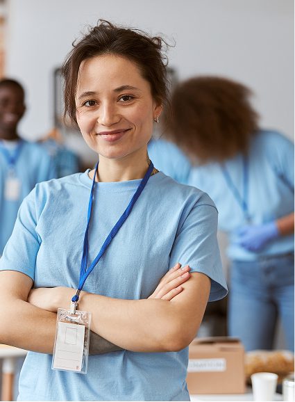 A young woman in a light blue T-shirt with a Sunflower Children's Hospice lanyard and name badge stands smiling with arms folded. Two people work in the background, also wearing blue shirts, in a well-lit indoor setting.