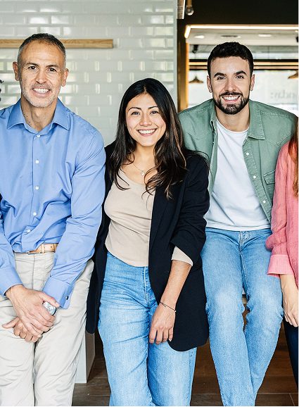 Three people smile at the camera indoors. A man in a blue shirt sits to the left, while a woman in a black blazer and jeans, representing Sunflower Children's Hospice, stands centre with a man in a green jacket and jeans on the right.