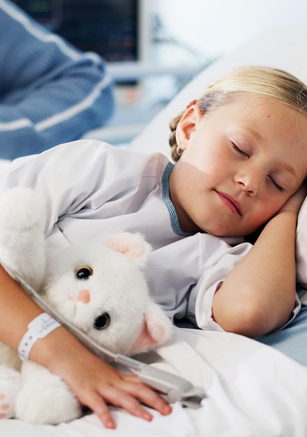 A young girl with blonde hair sleeps peacefully in a hospital bed at Sunflower Children’s Hospice, hugging a white cuddly cat. She wears a hospital wristband, and medical equipment is visible in the background.