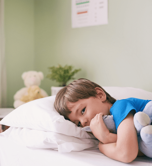 A young child in a blue shirt lies on a bed at Sunflower Children’s Hospice, resting their head on a white pillow and cuddling a soft toy. A blurred teddy bear and plant are visible in the background.