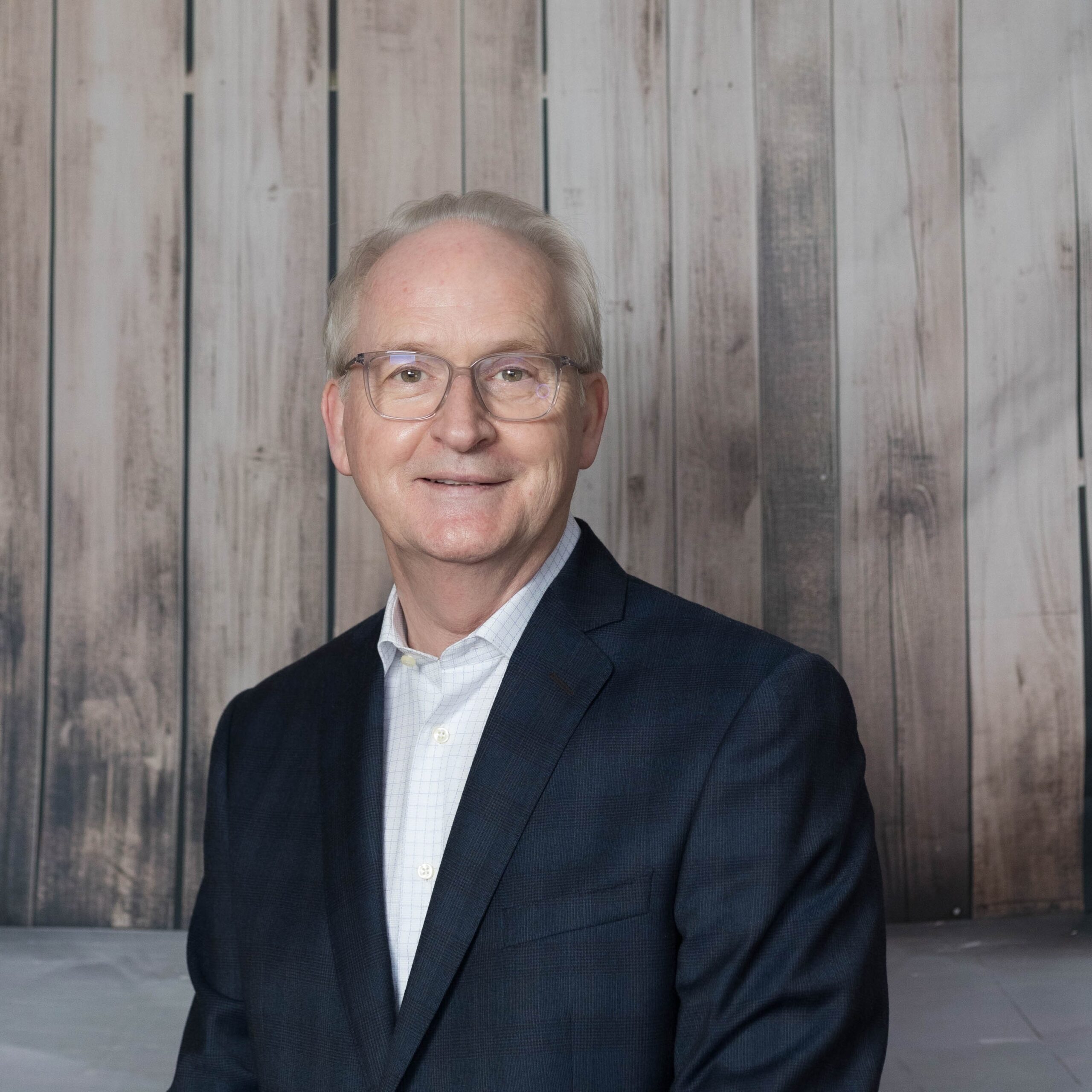 A smiling older man with short grey hair and glasses, wearing a dark suit and light-coloured shirt, stands in front of a wooden panel background at the Sunflower Children’s Hospice.
