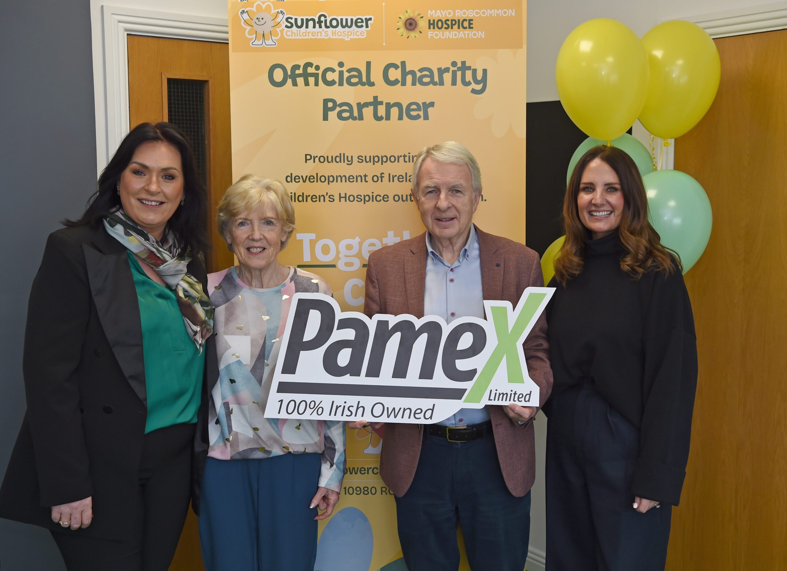 Four people stand smiling in front of a Sunflower Children's Hospice Charity Partnership banner, holding a Pamex sign. Yellow and green balloons decorate the right side, suggesting an uplifting indoor event atmosphere.