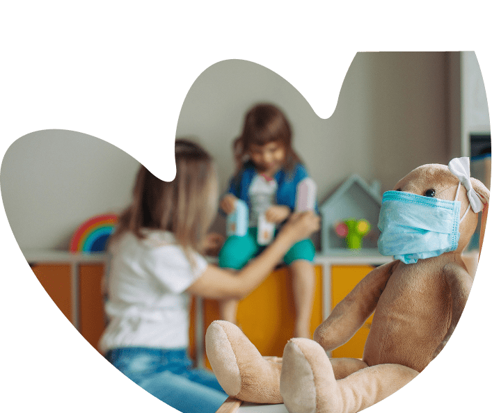 A stuffed toy wearing a face mask sits in the foreground, representing Sunflower Children’s Hospice, while two children play with toys in the blurred background. The image is framed with a rounded, abstract shape.