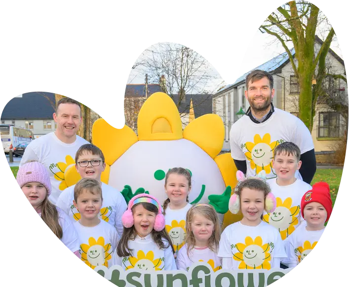 A group of children and two adults, all wearing white shirts with a yellow sunflower logo from Sunflower Children’s Hospice, pose and smile outside with a large sunflower mascot. The children wear colourful hats and earmuffs. Trees and buildings are in the background.