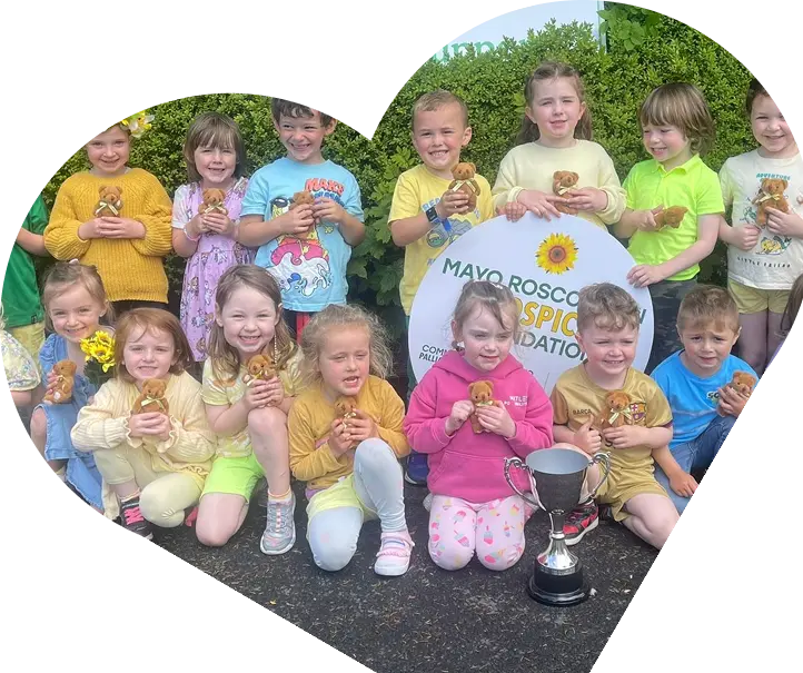 A group of children dressed in yellow and green pose outdoors, smiling and holding small teddy bears. They gather around a Mayo Roscommon Hospice Foundation sign and silver trophy, celebrating with Sunflower Children’s Hospice in a lush, green setting.