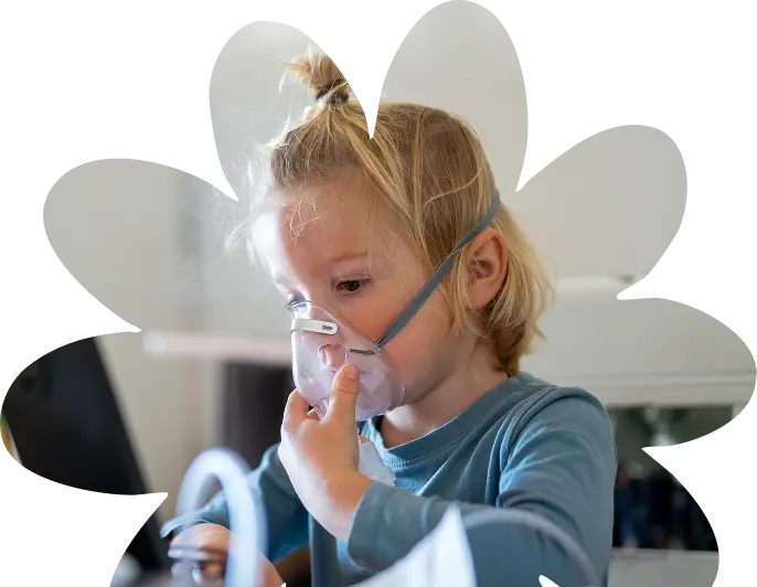 A young child with blonde hair wearing a blue shirt, supported by Sunflower Children’s Hospice, uses an oxygen mask whilst sitting at a table, looking intently at a laptop screen.