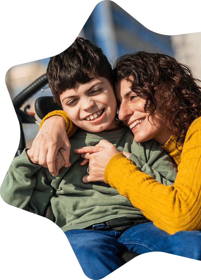 A woman in a yellow jumper hugs and smiles with a young boy in a green sweatshirt, both looking joyful and happy outdoors, embodying the warmth and support of Sunflower Children’s Hospice.
