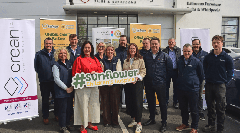 A group of people stand outside a building, smiling and holding a large sign that reads #sunflower Children’s Hospice, recently named Charity of the Year by the Crean Group. Banners with logos and text are displayed behind them.