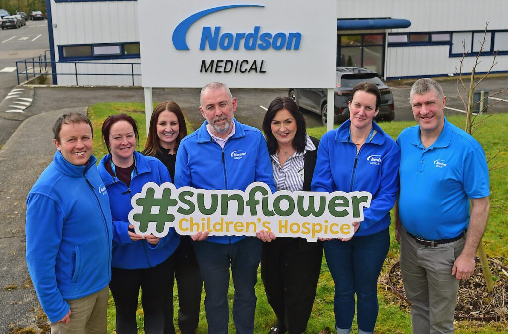 Seven people in Nordson Medical jackets stand outside the company building, smiling and holding a sign that reads #Sunflower Children’s Hospice, highlighting the partnership between Nordson Medical and Sunflower Children’s Hospice.
