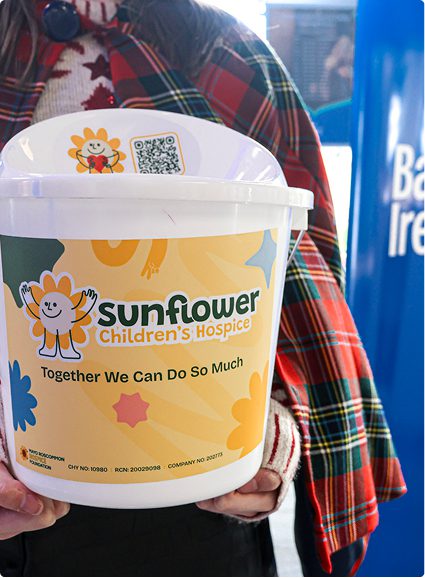 A person wearing a checked coat holds a donation bucket labelled Sunflower Children's Hospice with the slogan Together We Can Do So Much, featuring cheerful sunflower graphics.