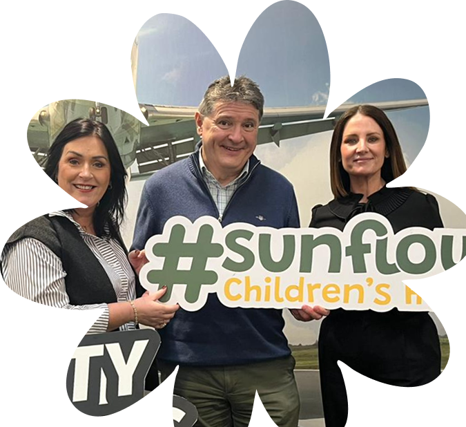 Three adults stand together smiling, holding a large Sunflower Children’s Hospice sign. They are in front of a backdrop with an aeroplane and blue sky visible.