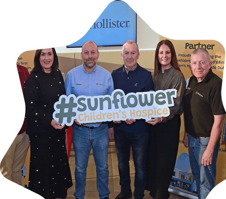 Six adults stand together smiling and holding a large sign that reads #sunflower Children’s Hospice. They are posing indoors with promotional banners in the background, celebrating corporate philanthropy and community impact.