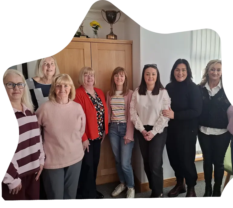 Eight women stand indoors in front of a wooden cupboard with a trophy and flowers on top at Sunflower Children’s Hospice. Smiling and dressed in casual clothing, they are bathed in sunlight streaming through a window on the right.