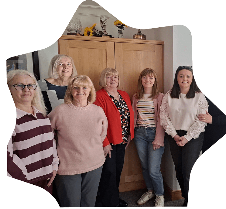 Six women stand together indoors in front of a wooden cupboard, smiling at the camera. Most are dressed casually, some wearing glasses. A vase with sunflowers, symbolising Sunflower Children’s Hospice, sits atop the cupboard.