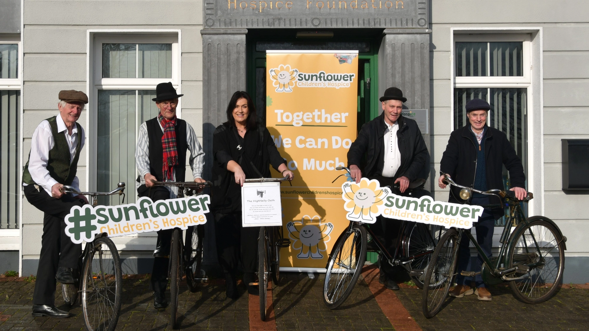 Five people stand with vintage High Nelly bicycles in front of a building, smiling. Two hold #sunflower Children's Hospice signs, and a sunflower banner behind them reads, Together We Can Do So Much—ready for the Charity Cycle.