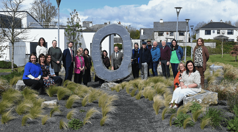 A group of eighteen people pose outdoors around a large circular stone sculpture in the Roscommon palliative care children’s memorial garden, set amidst landscaped grounds with modern buildings and a cloudy sky in the background.