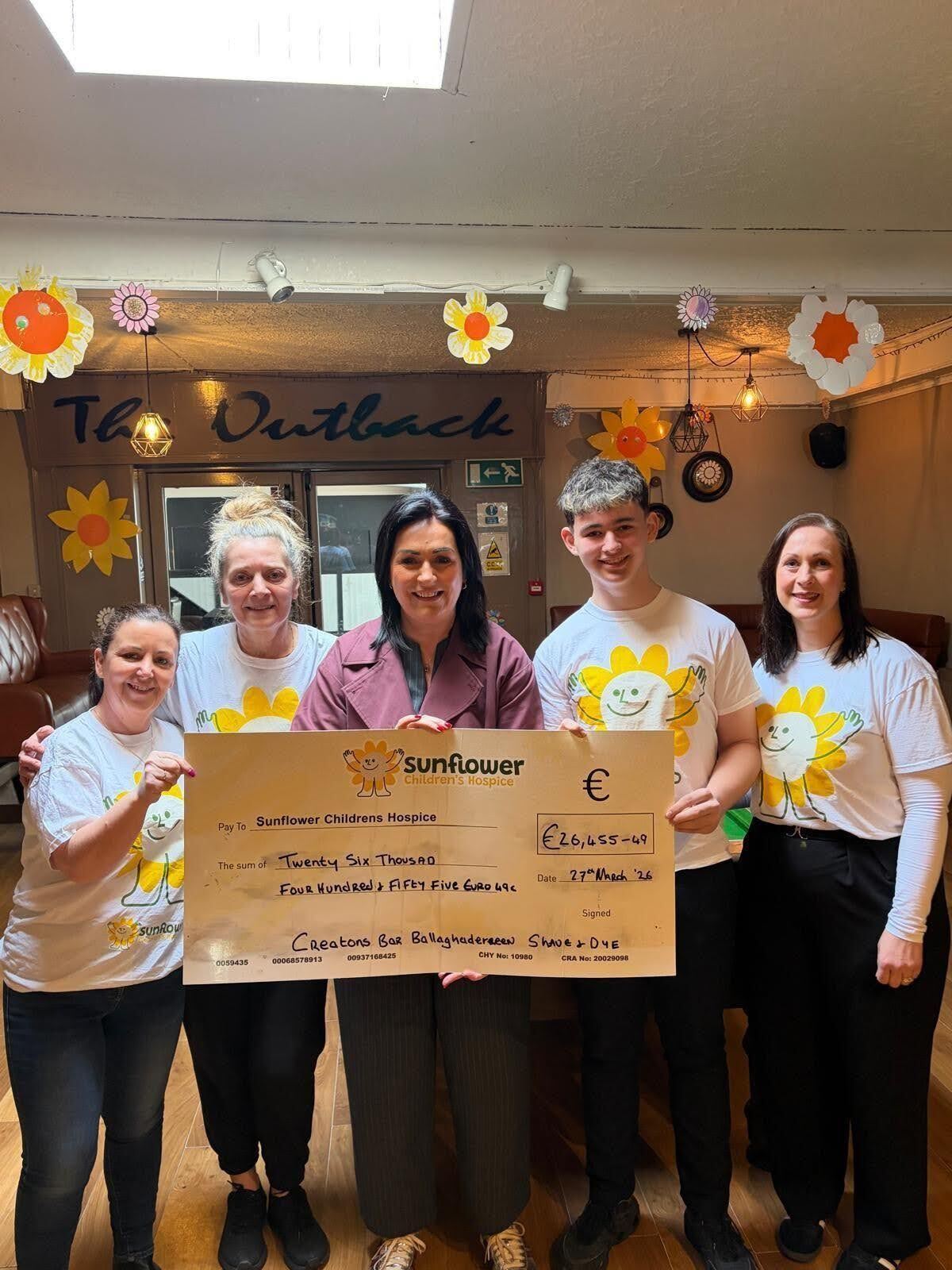 Five people stand indoors holding a large cheque for Sunflower Children's Hospice, celebrating a fundraising effort. Three wear white T-shirts with a smiling sunflower graphic. Flower decorations hang on the wall behind them.