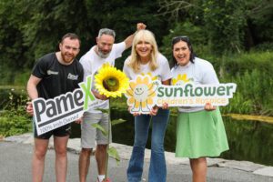 Four adults stand outdoors by a pond, smiling and holding signs for Pamex and Sunflower Children’s Hospice as part of the Linking Little Lives Campaign. A large artificial sunflower is in front of them, symbolising their fundraising efforts. Trees and greenery grace the background.
