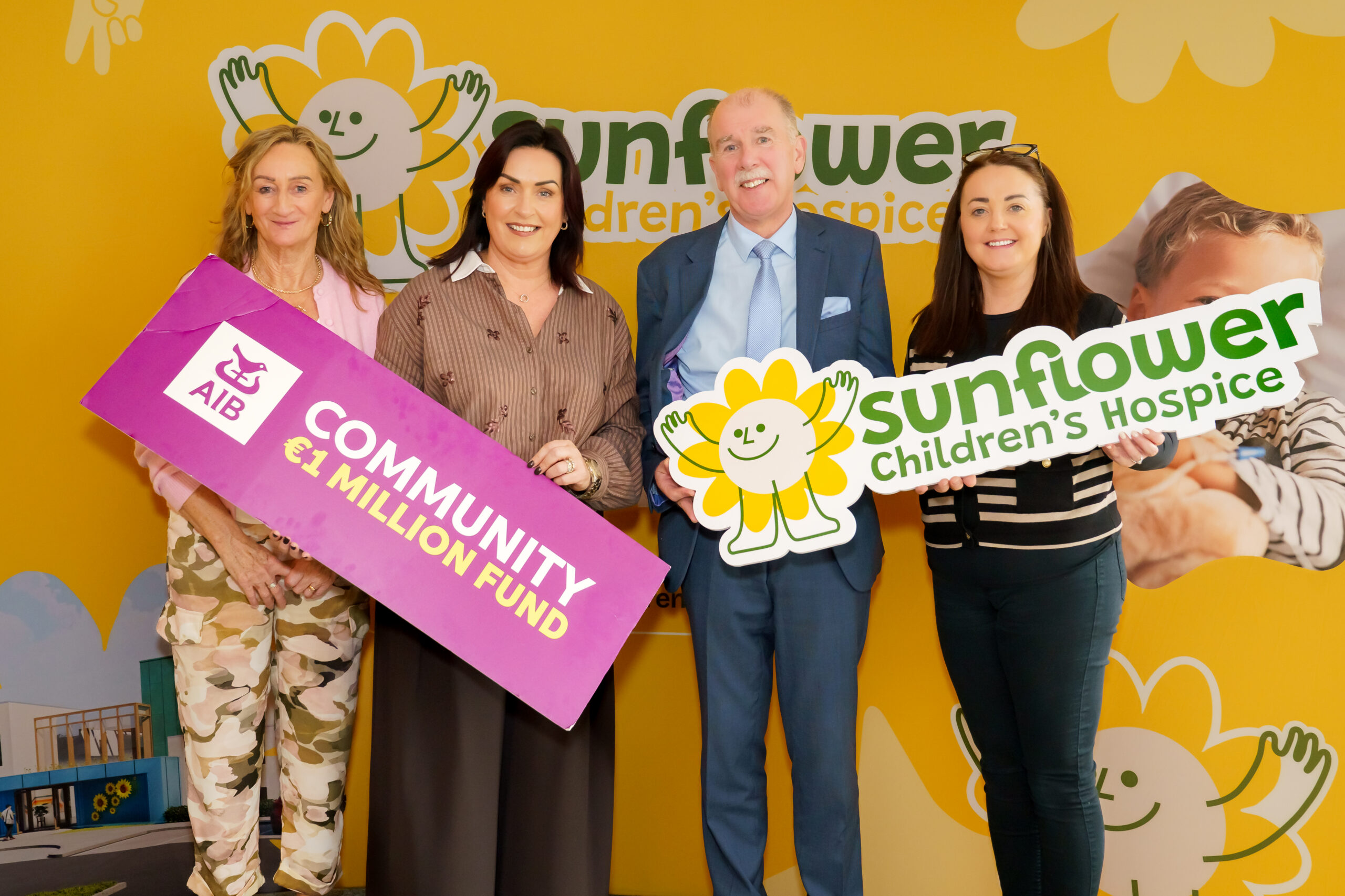 Four people stand in front of a bright yellow Sunflower Children’s Hospice backdrop, holding signs for AIB Community Fund and the hospice, showing their local support and smiling at the camera.